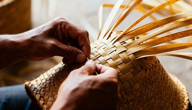 Artisan hands weaving straw hat with reed strips closeup showing woven texture and craft