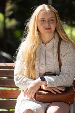Sunlit young woman sitting on park bench wearing knit cardigan holding leather bag