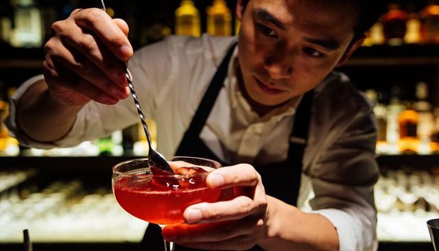 Stirring craft cocktail in coupe glass with bar spoon at low-lit bar mixologist at work