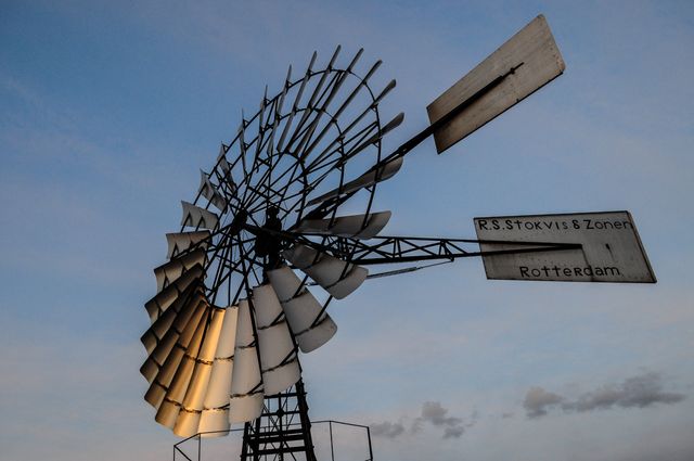 Historic windmill standing against a backdrop of the twilight sky. Silhouette effect highlights intricate metal structure and blades. Windmill inscription mentions 'R.S. Stokvis & Zonen' and 'Rotterdam'. Ideal for use in content relating to renewable energy, sustainability, historical architecture, and Dutch landmarks.