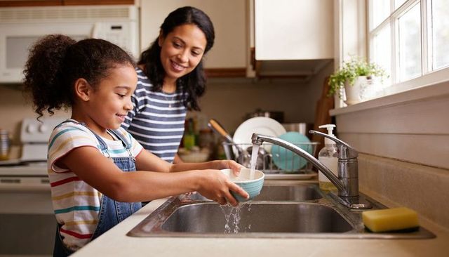 Mother teaching daughter to rinse bowl at sunlit home kitchen - family bonding chores