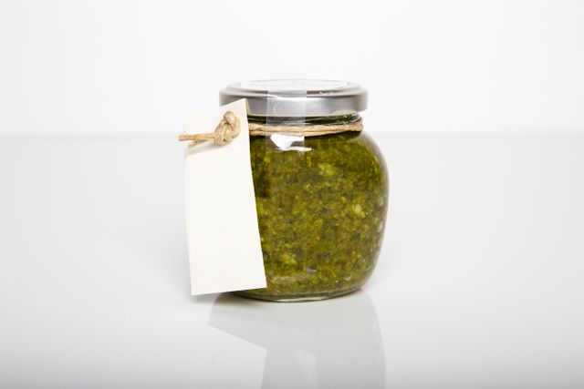 A clear glass jar filled with green homemade pesto, topped with a metal lid, and adorned with a blank tag attached with twine ribbon. This image is perfect for websites or blogs discussing homemade recipes, organic food, or cooking ingredients. It can also be used for marketing and branding of food products or to illustrate DIY gift ideas.