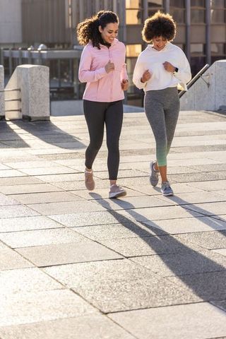 Female friends walking together in sunlit urban plaza wearing leggings and running shoes
