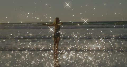 Blonde woman standing with arms outstretched in shallow surf at sunset with sparkling bokeh