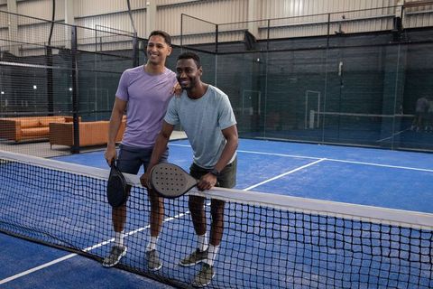 Smiling diverse male friends leaning on net holding padel rackets after indoor match on blue court