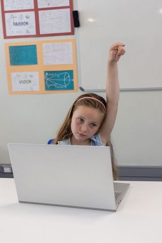Young girl raising hand while using laptop in classroom digital learning and participation
