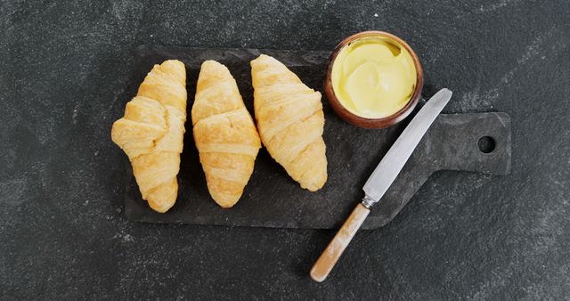 Freshly baked croissants are arranged on a slate board next to a jar of butter and a knife, with copy space. A top-down view captures the appeal of a simple yet indulgent breakfast or snack option.