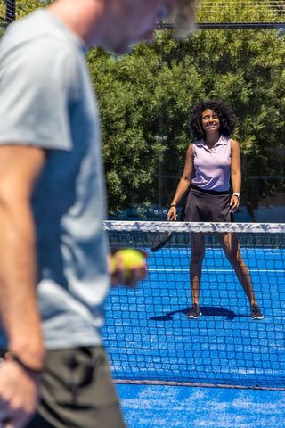 Sunlit mixed paddle match on blue court with smiling woman returning serve at net