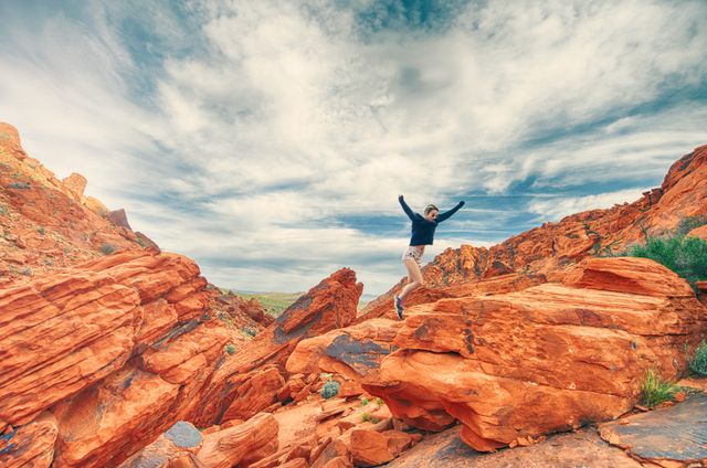 Person jumping on red rock formations against a beautiful sky with dynamic clouds. A perfect representation of adventure and freedom, suitable for promotions around traveling, tourism, outdoor activities, and nature exploring. This scenic landscape highlights desert beauty and excitement.