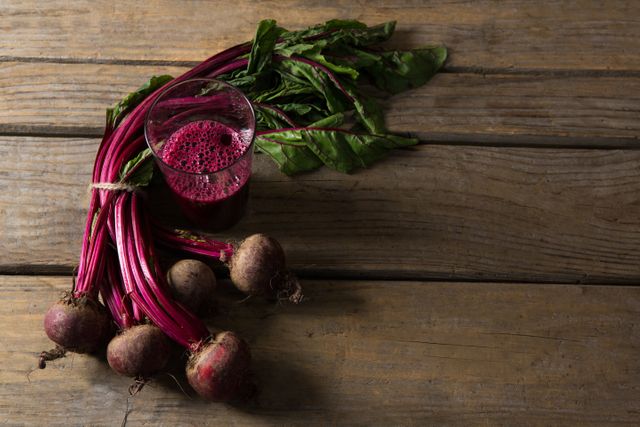 This image shows fresh beetroot with leaves and a glass of beetroot juice on a rustic wooden table. Ideal for use in health and wellness blogs, organic food promotions, farm-to-table restaurant advertisements, and nutritional guides. The earthy tones and natural setting emphasize the organic and wholesome nature of the produce.