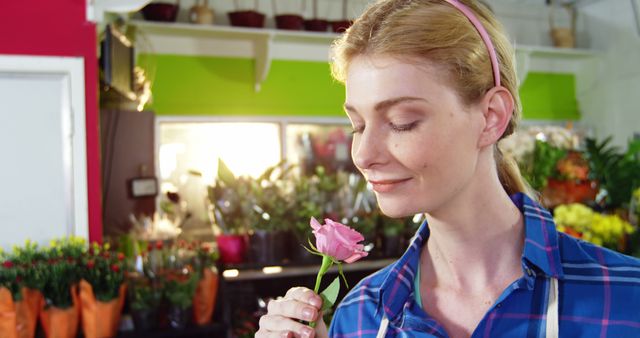 A young blonde woman smiling and enjoying the scent of a pink rose in a vibrant flower shop. Ideal for use in advertisements for florists, articles on relaxation and the benefits of aromatherapy, or promotional materials for flower-related products.
