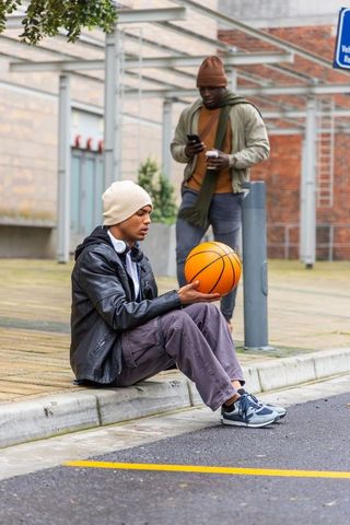 Diverse young men sitting on curb holding basketball checking smartphone urban street style