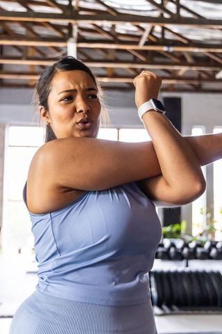Asian woman stretching upper body across chest in gym wearing activewear and smartwatch