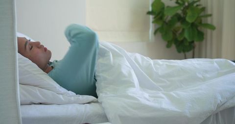 Mid-adult asian woman lying in bed under white duvet, wearing light blue top, minimalist calm bedroo