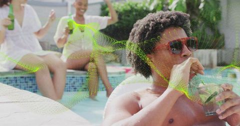 Shirtless man sipping mint lime cocktail at sunny backyard pool while friends lounge