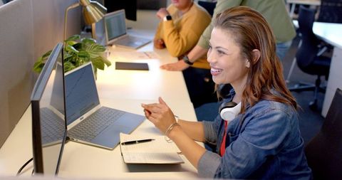 Smiling woman collaborating on video call in open-plan office with laptop and headset