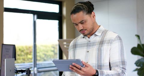 Asian man using tablet in modern office near large glass window, natural light workspace