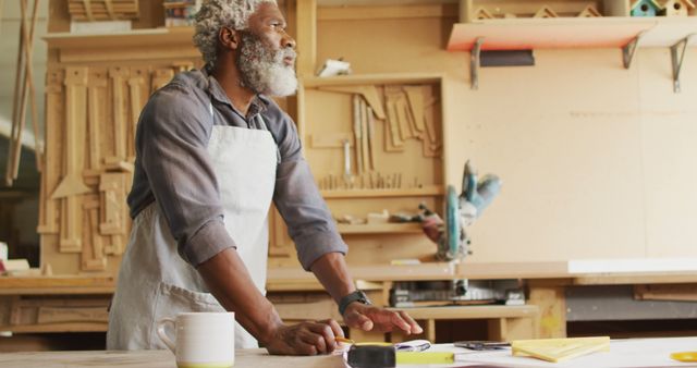 Senior woodworker standing at workbench planning new design. Suitable for themes related to craftsmanship, small business, creative work, and mature individuals in skilled trades. Ideal for advertisements promoting woodworking tools, carpentry services, or artisan craft projects.