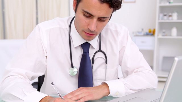 Male doctor clad in a formal shirt and tie, attentively writing on a clipboard, embodies professionalism and dedication. Modern medical office surroundings highlight a commitment to patient care. Useful for depicting healthcare practices, patient management scenes, or articles about medical professionalism.