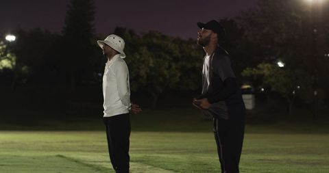 Two athletes standing on illuminated night field wearing bucket hat and baseball cap