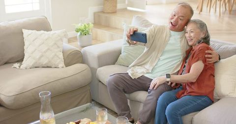 Senior asian couple taking selfie on cozy beige sofa in modern living room