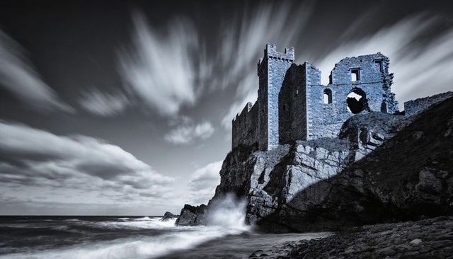 Storm-battered castle ruins towering on coastal cliff with crashing waves, moody monochrome