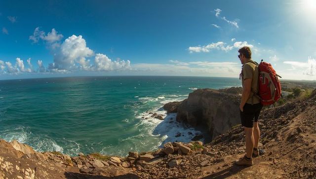 Solo hiker standing on rugged coastal cliff surveying turquoise ocean with red backpack