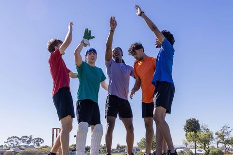 Male cricket teammates celebrating victory on sunny field raising hands for high-five