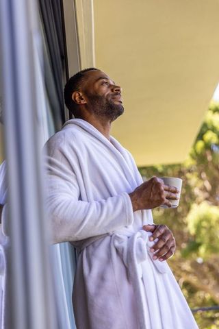 African american man enjoying coffee on balcony in white bathrobe, serene morning