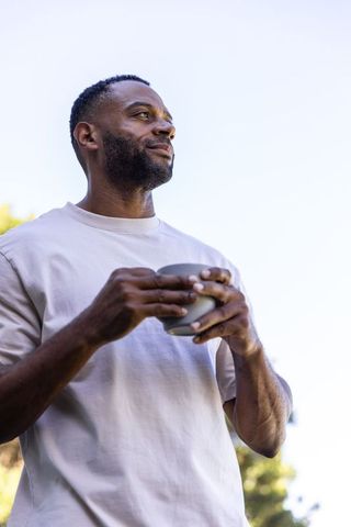 Sunlit african american man standing in garden holding ceramic mug, casual morning