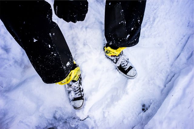 Snow-covered ground, person wearing yellow bands by ankles, classic Converse sneakers. Useful for themes on winter, fashion, casual wear, cold weather, and outdoor activities in snowy conditions.