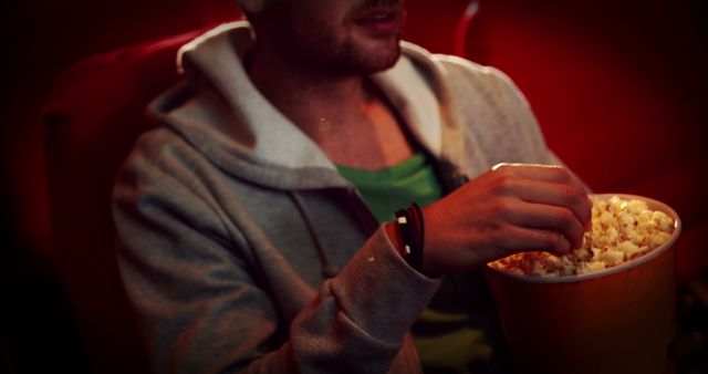 Man enjoying popcorn in dark cinema while watching a movie. Wearing casual clothing, seated comfortably. Ideal for illustrating cinema experiences, entertainment activities, and leisure lifestyle. Can be used in ads, blogs, and articles related to movies, theaters, or popcorn snacks.