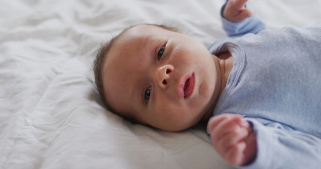 Newborn baby wearing a blue onesie is lying down on a white bed, appearing calm and serene. Perfect for use in parenting blogs, baby product advertisements, family newsletters, and childcare educational materials.