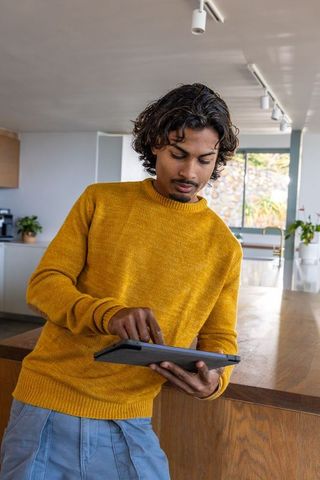 Asian man tapping tablet at modern kitchen island wearing mustard sweater and blue pants