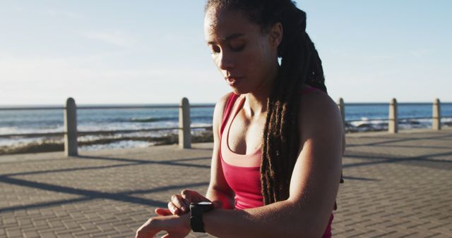A fit woman with dreadlocks is checking her smartwatch during an outdoor workout near the ocean. She is in sportswear and appears focused on her fitness routine. This image is ideal for use in blogs, fitness websites, health and wellness articles, advertisements promoting active lifestyles, or smartwatch and athletic wear promotions.