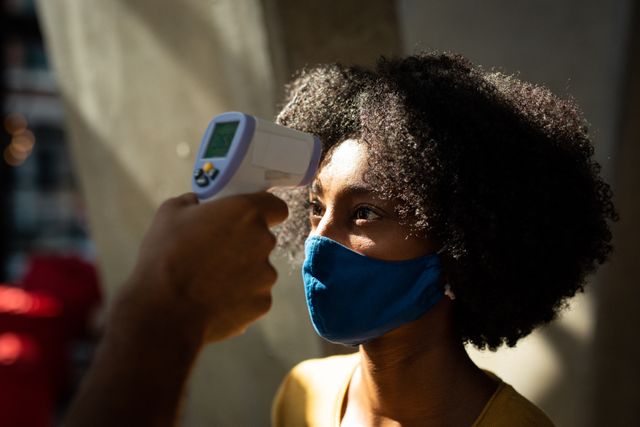 African American woman wearing face mask having temperature taken by colleague before work. Useful for illustrating workplace safety measures during the COVID-19 pandemic, health and safety protocols, and preventive actions in business environments.