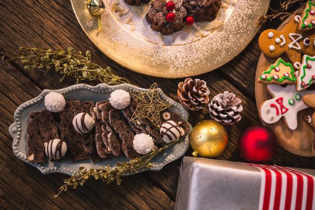 Close-up of various Christmas desserts and gifts on a wooden table. The image features an assortment of cookies, chocolate treats, and festive decorations such as pinecones and ornaments. Ideal for holiday-themed marketing, social media posts, and festive greeting cards.