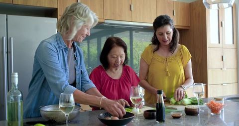Mature women cooking together at kitchen island, chopping vegetables and enjoying wine