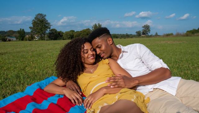 Expecting couple relaxing on meadow touching pregnant belly on picnic blanket