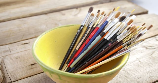 Assorted paint brushes standing in a yellow bowl on a rustic wooden table. Ideal for articles or advertisements related to arts and crafts, painting tutorials, creative workshops, or art supply stores.