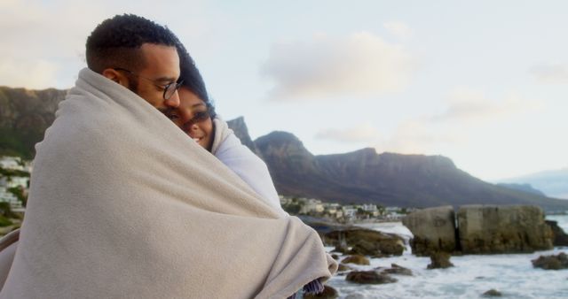 Romantic diverse couple with blankets on backs standing on beach and embracing, copy space. Summer, vacation, romance, love, relationship, free time and lifestyle, unaltered.