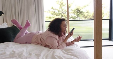 Young woman lying on bed speaking into phone wearing pink hoodie and socks by balcony