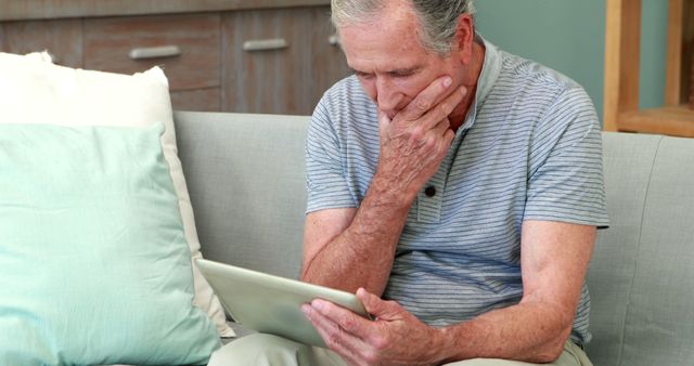 Senior man intently using tablet while sitting on sofa at home. Suitable for themes related to technology adoption by older adults, senior living, leisure activities at home, and digital connectivity among the elderly.