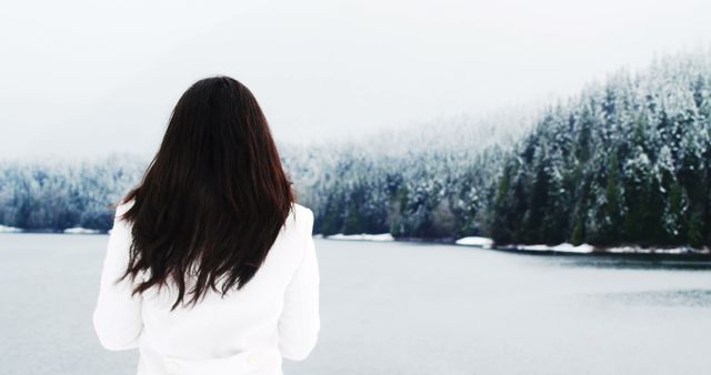 Woman in white coat is standing near a snow-covered forest and calm lake, viewing the natural winter scene. Ideal for use in content about nature, peaceful moments, winter adventure, introspective themes, and travel destinations.