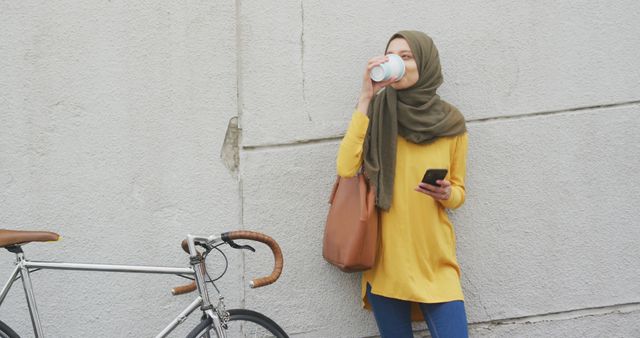 Happy biracial woman in hijab standing by bike in city street drinking coffee and using smartphone. City living, communication, transport and modern urban lifestyle.