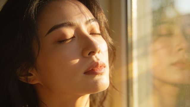 Asian woman basking in golden sunlight by window, eyes closed, reflective glass portrait