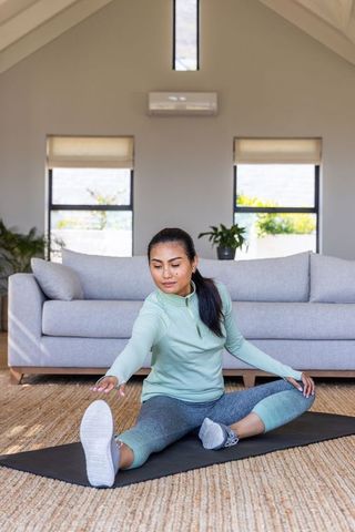 Asian woman performing seated hamstring stretch on yoga mat in bright living room for home fitness