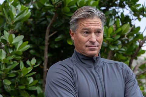 Thoughtful middle-aged man wearing gray zip-up jacket standing by green shrubs looking left