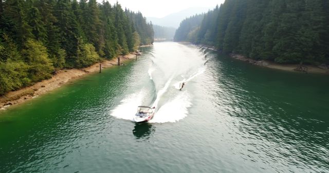 Scene captures an aerial view of a speedboat towing a waterskier on calm lake waters, flanked by dense forest on both sides. Ideal for depicting outdoor leisure, summertime activities, water sports, nature exploration, travel destinations, and adventure themes.