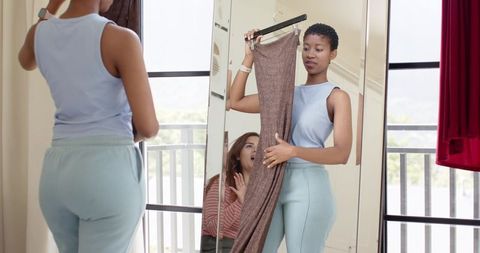 Young women trying on brown dress by balcony mirror, casual styling and wardrobe advice